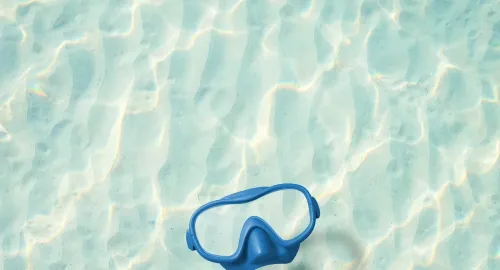 Maschera blu sospesa su sabbia chiara, riflessi d'acqua creano un'atmosfera serena.